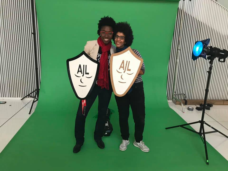 Timnit Gebru and Joy Buolamwini stand together against a plain backdrop, holding Algorithmic Justice League shields and smiling.